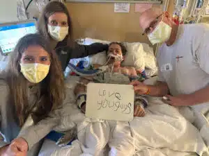 Logan - laying in PICU bed, surrounded by family holding a message board with, "Love you guys"