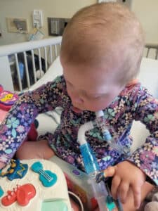 photo of a little girl in a flower print shirt with a trach in a hospital bed playing with a sound toy