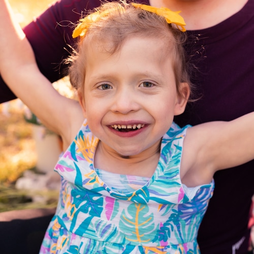 photo of a girl outside with her arms up while smiling and her mother in the background